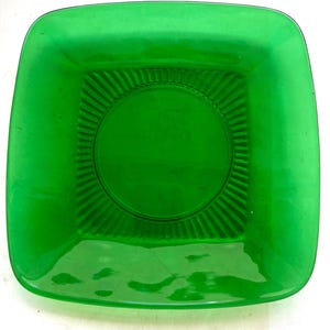Peut inclure: Une assiette carrée en verre vert émeraude. L'assiette a des coins arrondis et un motif circulaire légèrement surélevé au centre, entouré de lignes radiales. La surface de l'assiette est brillante, reflétant la lumière.