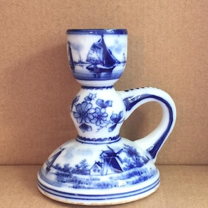 Puede incluir: Un candelero de cerámica azul y blanco con asa. El candelero presenta una escena holandesa con un molino de viento, casas y un velero.