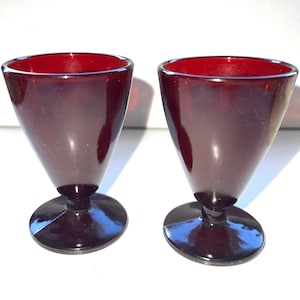 May include: Two ruby red glass goblets with a conical bowl and a circular base. The glasses have a glossy finish and are set against a white background. The stemware is likely for serving beverages.
