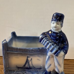 Puede incluir: Macetero de cerámica azul y blanco con forma de niño holandés que sostiene un acordeón. El macetero tiene un pequeño diseño de velero en el lateral.