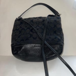May include: Black leather and canvas shoulder bag with a signature Coach pattern. The bag has a long adjustable strap and a top zipper closure.