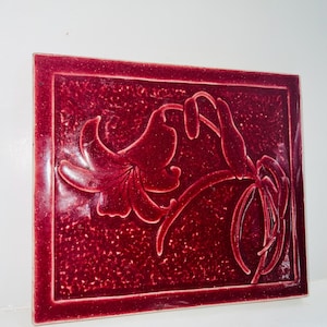 May include: A red ceramic tile with a raised design of a stylized flower. The tile has a textured surface and a border.