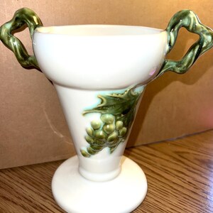 May include: White ceramic vase with a flared top and a round base. The vase features green, vine-like handles and a grape cluster design. The handles and grape design have a glossy finish.