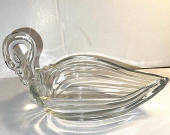 May include: Clear glass swan-shaped dish with a curved neck and a wide, ribbed body. The dish is designed with a graceful, flowing form, and the glass has a slight iridescence. The dish is likely used for serving or display.