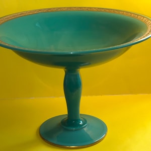 Puede incluir: Un cuenco de pedestal de vidrio verde azulado con un borde dorado. El cuenco está sobre un fondo amarillo.
