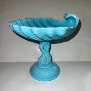 Puede incluir: Un plato de vidrio azul claro en forma de concha de mar con un pie en forma de cola de sirena. El plato es perfecto para servir dulces o pequeñas chucherías.