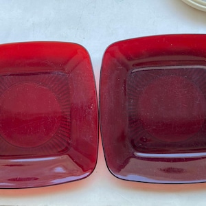 May include: Two square, ruby red glass plates. Each plate has a slightly rounded square shape with a raised, circular design in the centre. The plates are a deep, solid red colour, and appear to be vintage or antique.