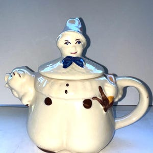 Pode incluir: Uma chaleira de cerâmica vintage em forma de boneco de neve. A chaleira é principalmente branca com detalhes em marrom, uma gravata borboleta azul e um chapéu azul. O bico é uma cabeça de porco e a alça é um laço curvo.