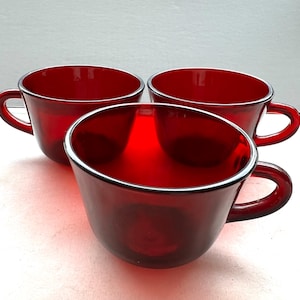 May include: Three ruby red glass teacups with rounded handles. The cups have a simple, elegant design with a silver rim around the top. The cups are arranged in a group, showcasing their rich color and classic shape.