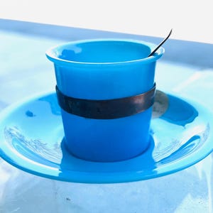 May include: A vibrant blue teacup and saucer. The cup features a dark band and a small spoon. The saucer, in a matching blue, has a decorative pattern. The set rests on a light blue surface, creating a cohesive look.