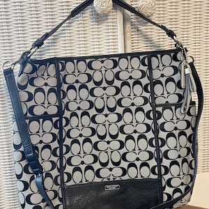 May include: Black and gray tote bag with a repeating pattern of interlocking Cs. The bag has black leather accents, a shoulder strap, and a small leather pocket with a silver logo. A silver chain and tassel hang from the side.