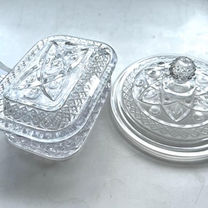 Peut inclure: Deux plats de service en verre taillé transparent. L'un est rectangulaire avec un couvercle et une cuillère, l'autre est rond avec un bouton décoratif sur le couvercle. Les deux présentent des motifs géométriques complexes.