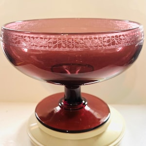 Puede incluir: Un cuenco de vidrio rojo oscuro con un diseño blanco grabado alrededor del borde. El cuenco se asienta sobre un pedestal rojo a juego con una base blanca.