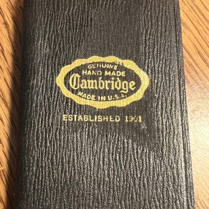 May include: Black, textured, rectangular wallet with the Cambridge logo in gold. The logo reads "Genuine Hand Made Cambridge Made in U.S.A." and "Established 1901".
