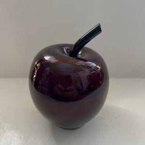 Può includere: Una mela di vetro di colore viola scuro con un gambo nero. La mela è lucida e ha una superficie liscia.