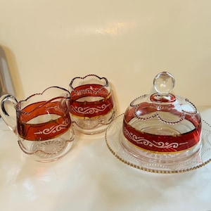 Puede incluir: Un juego de mantequillera de vidrio vintage con un diseño rojo y dorado. El juego incluye una mantequillera con tapa, una jarra de crema y un azucarero.