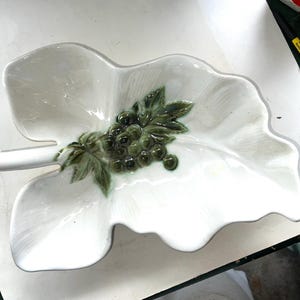 May include: White ceramic leaf-shaped serving dish with a raised green grape cluster design. The dish has a glossy finish and a stem-like handle. Suitable for serving appetizers or as a decorative piece.