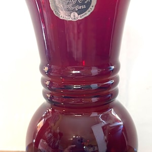 May include: A ruby red glass vase with a bulbous base and a flared top. The vase has a series of horizontal ridges around the middle. A silver and white label reads "Royal Ruby Anchorglass" and "Lancaster, Ohio, U.S.A."