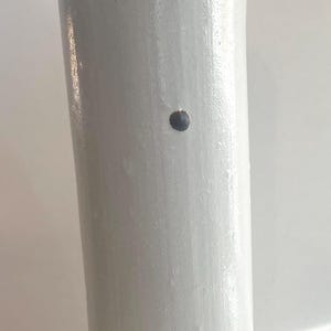 May include: A tall, off-white ceramic bottle with a narrow neck and a cylindrical body. The bottle has a dark spot and a small embossed design near the base. The bottle is approximately 12 inches tall.