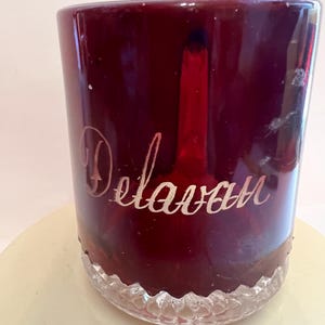 以下が含まれることがあります： 筆記体で「Delavan」の文字が刻まれた、バーガンディ色のグラス。 グラスの底には装飾的でテクスチャ加工されたデザインがあります。 グラスは円筒形で、クリーム色の表面に置かれています。