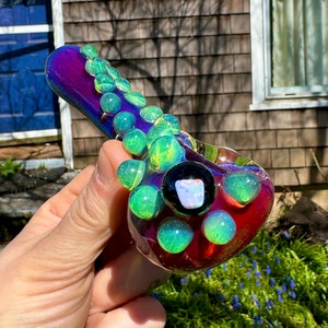 May include: A glass pipe with a purple, red, and green iridescent finish. The pipe has a series of green glass bubbles along the side and a round bowl with a black and white opal inlay.