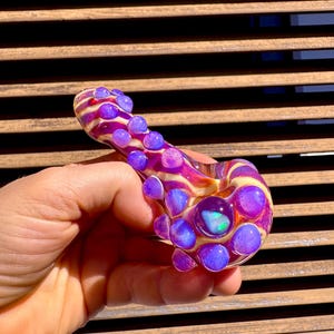 Puede incluir: Una pipa de vidrio soplado a mano con un patrón arremolinado de amarillo, rosa y morado. La pipa está adornada con múltiples burbujas de vidrio morado y una gran piedra central iridiscente. El diseño es artístico y colorido.
