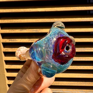May include: A colorful glass pipe with a fish-like design. The pipe features shades of blue, red, and clear glass, with a textured, scale-like pattern. The eye is a deep red, and the overall shape is rounded and organic.