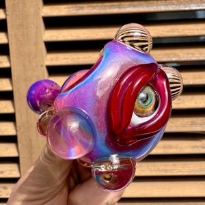 May include: A colorful glass art piece with a unique design. The piece features a combination of purple, blue, and red hues, with a prominent eye-like shape. It has multiple spherical and rounded elements, and striped orbs. The glass appears iridescent.