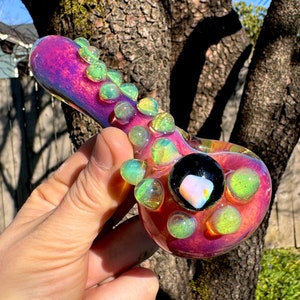 May include: A glass pipe with a pink iridescent finish and green and yellow accents. The pipe has a round bowl with a small, clear, iridescent stone in the center.