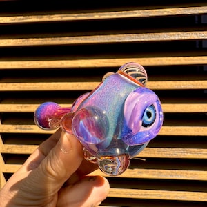 Puede incluir: Una pipa de vidrio soplado a mano con forma de pez. Presenta tonos de azul, morado y rosa, con una base transparente y redonda y un ojo detallado. El diseño incluye una boquilla pequeña y esférica y un cuenco más grande y redondeado.