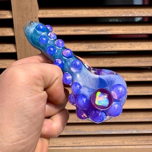 Puede incluir: Una pipa de vidrio azul y morado con un diseño bulboso. La pipa presenta un cuenco grande y redondo con un centro iridiscente y está adornada con múltiples adornos esféricos pequeños. El vidrio tiene una apariencia veteada y arremolinada.