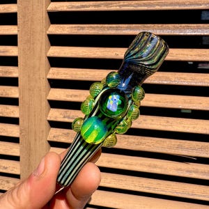 May include: A glass pipe featuring a black and light green striped pattern. The body is adorned with several small, raised green glass spheres and a larger, central green glass marble with an iridescent, multi-colored reflective finish.