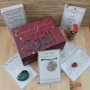 May include: A wooden box with the words "Come Gather for a Spell" on the lid. The box is surrounded by various items, including a small jar with a red lid, a card with the words "Love Blessing Jar", a card with the words "The Wild Place", a card with the words "Earth Witch", a card with the words "Moonstone", a card with the words "Red Jasper", and a small painted rock with a moon and trees.