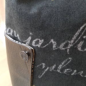 May include: Close-up of a black canvas tote bag with a brown leather trim and a metal rivet. The bag has a white inscription that reads "au jardin" and "yellow".