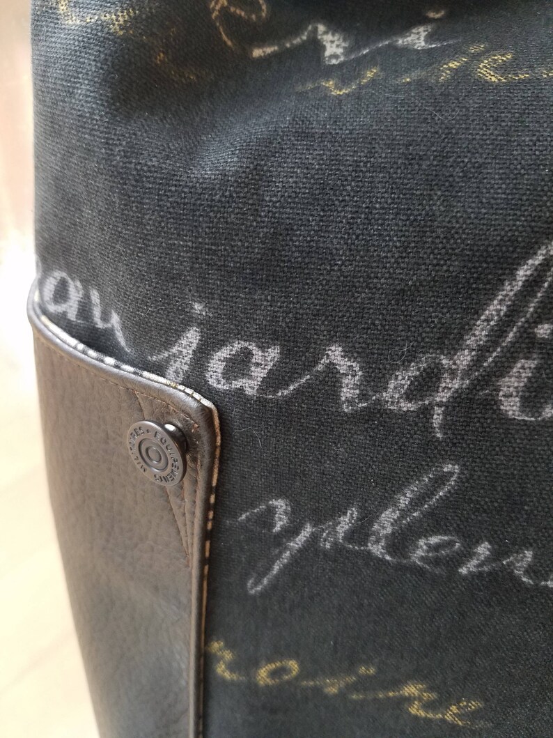 May include: Close-up of a black canvas tote bag with a brown leather trim and a metal rivet. The bag has a white inscription that reads "au jardin" and "yellow".