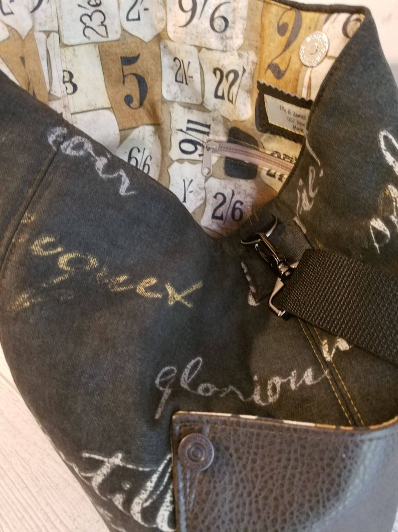 May include: A black canvas tote bag with a brown leather base and a zipped pocket. The bag has a black strap and is decorated with white and gold writing. The inside of the bag is lined with a fabric featuring a pattern of numbers and a zipped pocket.