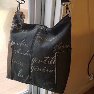 May include: A black canvas shoulder bag with a brown leather flap and a strap. The bag has a gold and white print of French words on it.