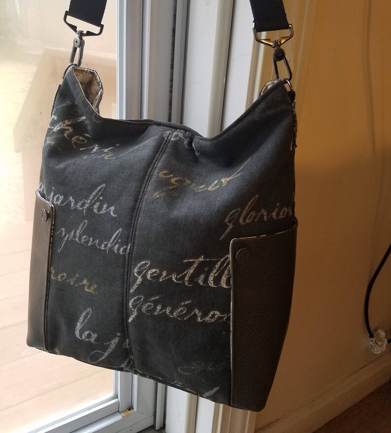 May include: A black canvas shoulder bag with a brown leather flap and a strap. The bag has a gold and white print of French words on it.
