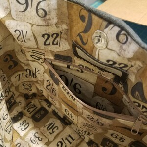 May include: A close-up of the inside of a tote bag with a zipped pocket. The fabric is brown with a pattern of numbers and fractions. The zipper is brown and the zipper pull is silver.