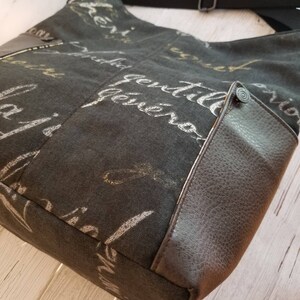 May include: A black canvas tote bag with a brown leather flap and a silver zip. The bag has a white and gold print of cursive writing on the front.