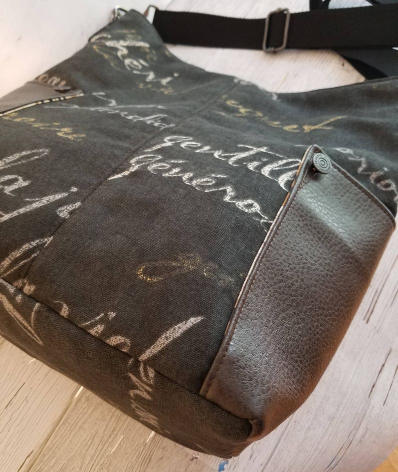 May include: A black canvas tote bag with a brown leather flap and a silver zip. The bag has a white and gold print of cursive writing on the front.