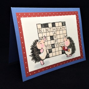 Op de afbeelding: Een kaart met een rode en witte stippenrand. Twee egels werken aan een kruiswoordpuzzel met de tekst "Happy Birthday to You".