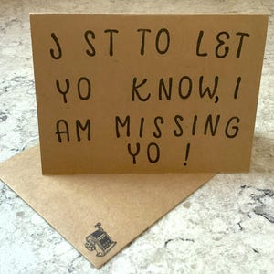 May include: A brown greeting card with the message "J ST TO L&T YO KNOW, I AM MISSING YO!" in black lettering. The card is on top of a matching envelope with a small illustration of a machine.