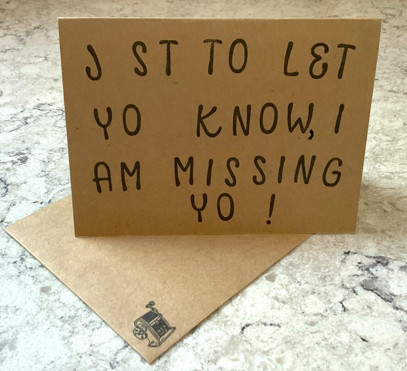 Puede incluir: Tarjeta de felicitaci&oacute;n marr&oacute;n con el mensaje "J ST TO L&T YO KNOW, I AM MISSING YO!" en letras negras. La tarjeta est&aacute; sobre un sobre a juego con una peque&ntilde;a ilustraci&oacute;n de una m&aacute;quina.