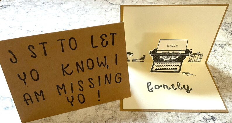 Puede incluir: Dos tarjetas de felicitaci&oacute;n. Una es marr&oacute;n con texto escrito a mano en negro: "J ST TO L&T YO KNOW, I AM MISSING YO!". La otra es una tarjeta desplegable con una ilustraci&oacute;n de m&aacute;quina de escribir y la palabra "hello" en el papel. La palabra "fontly" est&aacute; escrita debajo.