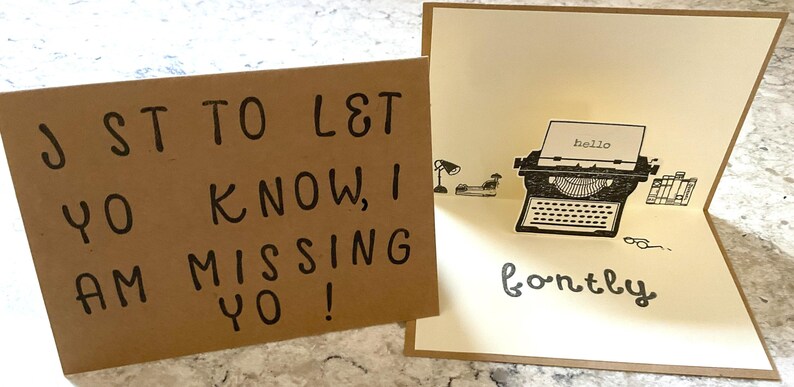 Puede incluir: Una tarjeta de felicitaci&oacute;n hecha a mano con un dise&ntilde;o de m&aacute;quina de escribir emergente. La tarjeta est&aacute; abierta, revelando una ilustraci&oacute;n de m&aacute;quina de escribir vintage con la palabra "hello" en el papel. La tarjeta dice "fontly" en la parte inferior. La tarjeta adjunta dice "J ST TO L& YO KNOW, I AM MISSING YO!"