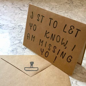 Puede incluir: Una tarjeta de felicitaci&oacute;n marr&oacute;n con el texto escrito a mano "JST TO LET YO KNOW, I AM MISSING YO!" est&aacute; apoyada, con un sobre a juego delante. El sobre tiene un dise&ntilde;o de sello.