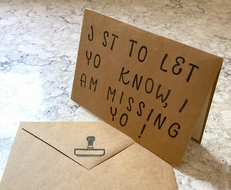 Puede incluir: Una tarjeta de felicitaci&oacute;n marr&oacute;n con el texto escrito a mano "JST TO LET YO KNOW, I AM MISSING YO!" est&aacute; apoyada, con un sobre a juego delante. El sobre tiene un dise&ntilde;o de sello.