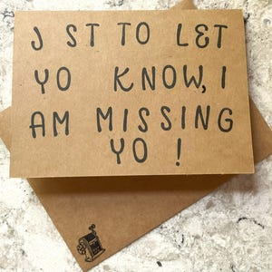 Puede incluir: Una tarjeta de felicitaci&oacute;n de papel kraft con el mensaje escrito a mano "J ST TO L&T YO KNOW, I AM MISSING YO!" en tinta negra. La tarjeta est&aacute; dentro de un sobre a juego con una peque&ntilde;a ilustraci&oacute;n de un sacapuntas.