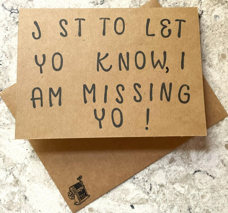 Puede incluir: Una tarjeta de felicitaci&oacute;n de papel kraft con el mensaje escrito a mano "J ST TO L&T YO KNOW, I AM MISSING YO!" en tinta negra. La tarjeta est&aacute; dentro de un sobre a juego con una peque&ntilde;a ilustraci&oacute;n de un sacapuntas.